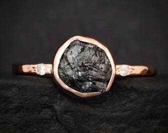 May include: A rose gold ring featuring a large, central, raw black stone with a rough, textured surface. The band is hammered and includes two small, clear, round-cut accent stones set on either side of the central setting in a bezel style.