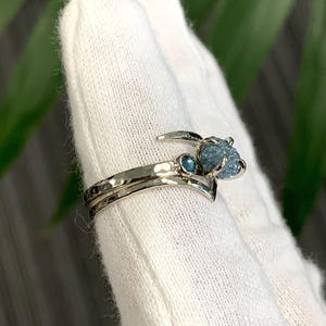 May include: A silver-toned ring featuring a hammered band with a V-shaped contour. The ring is set with a small, round, faceted blue gemstone and a larger, rough, uncut blue gemstone held in a prong setting at the top of the band.