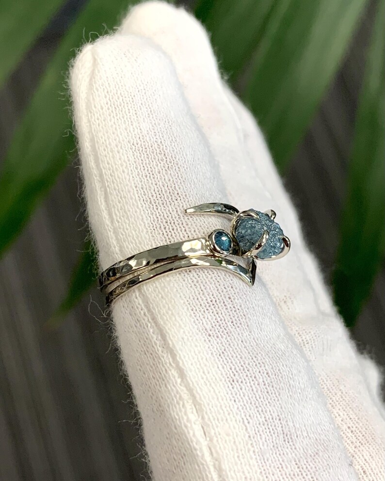May include: A silver-toned ring featuring a hammered band with a V-shaped contour. The ring is set with a small, round, faceted blue gemstone and a larger, rough, uncut blue gemstone held in a prong setting at the top of the band.