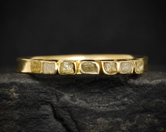 Può includere: Un anello a fascia in oro con una fila di sette piccoli diamanti grezzi di colore giallo pallido incastonati orizzontalmente sulla parte superiore. Il metallo ha una finitura liscia e lucida che contrasta con le pietre irregolari.