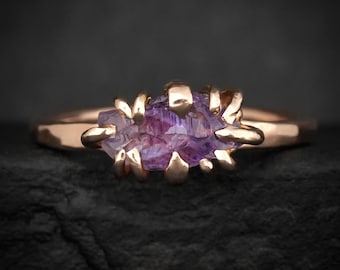 May include: A rose gold ring featuring a raw, elongated purple gemstone set in a claw-style prong setting. The band has a smooth, polished finish, and the central stone displays natural facets and deep violet hues held securely by multiple metal prongs.