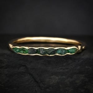 Op de afbeelding: Een gouden ring met een rij groene smaragden. De band heeft een gladde, gepolijste afwerking, terwijl de smaragden een ruwe, natuurlijke textuur hebben. De ring is geplaatst tegen een donkere achtergrond.