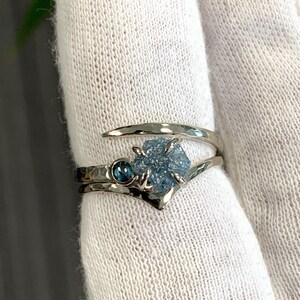May include: A silver-toned bypass ring featuring a hammered metal band. The ring is set with a central, rough-cut, light blue gemstone held by four prongs, accompanied by a smaller, round, dark blue faceted gemstone set directly beside it.