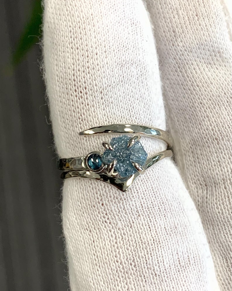 May include: A silver-toned bypass ring featuring a hammered metal band. The ring is set with a central, rough-cut, light blue gemstone held by four prongs, accompanied by a smaller, round, dark blue faceted gemstone set directly beside it.