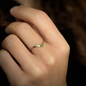 Può includere: Un delicato anello d'oro con una piccola pietra preziosa quadrata di colore verde smeraldo incastonata al centro. L'anello ha la forma di una V ed è indossato su un dito.