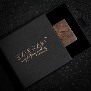 May include: A black square jewelry box with a sliding lid featuring the copper-colored text EMERAKI rough fine jewelry EST.2018. Inside the box, a small rectangular wooden piece with a geometric triangle logo is partially visible.