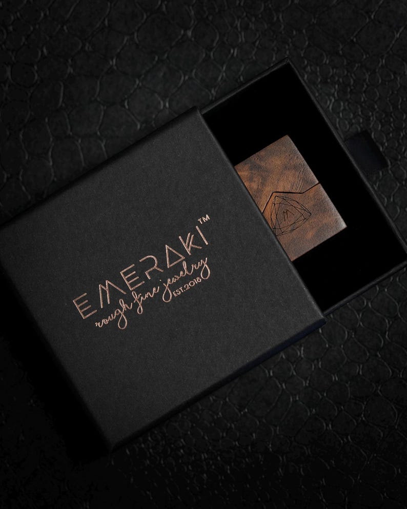 May include: A black square jewelry box with a sliding lid featuring the copper-colored text EMERAKI rough fine jewelry EST.2018. Inside the box, a small rectangular wooden piece with a geometric triangle logo is partially visible.