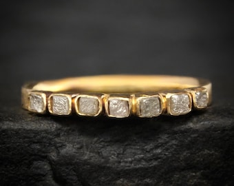 Può includere: Un anello a fascia in oro con sette piccole pietre bianche traslucide a taglio quadrato incastonate in fila sulla parte superiore. Il metallo ha una finitura gialla lucida e le pietre hanno un aspetto opaco e testurizzato, creando un contrasto con l'oro.