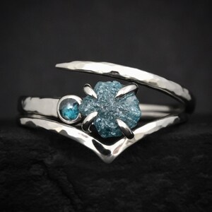 May include: A silver bypass ring featuring a hammered texture, a small round blue gemstone, and a larger raw, uncut blue diamond held in a four-prong setting. The band has a V-shaped design that curves around the central stones.