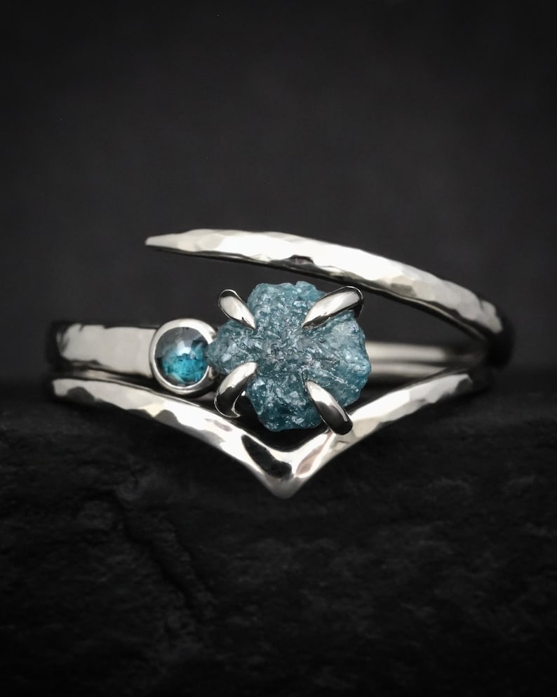 May include: A silver bypass ring featuring a hammered texture, a small round blue gemstone, and a larger raw, uncut blue diamond held in a four-prong setting. The band has a V-shaped design that curves around the central stones.