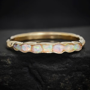 Op de afbeelding: Een gouden ring met een rij opalen. De opalen hebben een melkachtige witte basis met iriserende kleuren. De band van de ring heeft een getextureerd, gehamerd uiterlijk.