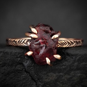 Può includere: Anello con una gemma grezza di colore rosso intenso. La gemma è tenuta in posizione da griffe color oro. La fascia dell'anello presenta un motivo intrecciato dettagliato. L'anello è posto su uno sfondo scuro.