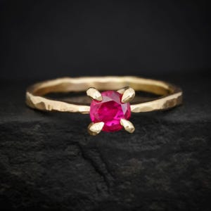 Könnte beinhalten: Ein goldener Ring mit gehämmertem Band und einem leuchtend roten, runden Edelstein. Der Edelstein wird von vier Krappen gehalten. Der Ring ist vor einem dunklen Hintergrund platziert.