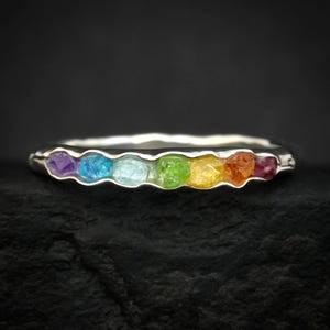 May include: A silver ring with a rainbow of gemstones. The ring features a series of colourful stones, including purple, blue, green, yellow, orange, and red. The ring is set against a dark background.