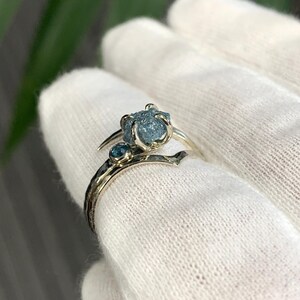 May include: A silver-toned ring featuring a prominent, rough-cut, light blue gemstone held in a prong setting, accompanied by a smaller, round, faceted blue stone set directly into the band, which has a textured, hammered metal finish.