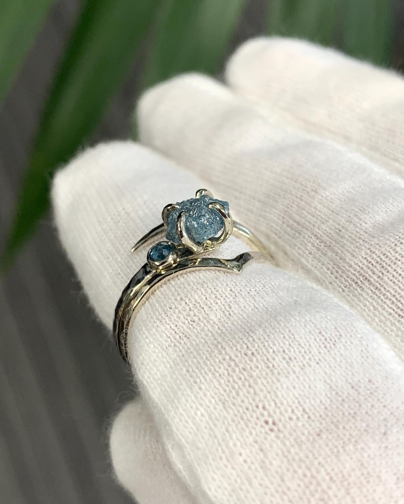 May include: A silver-toned ring featuring a prominent, rough-cut, light blue gemstone held in a prong setting, accompanied by a smaller, round, faceted blue stone set directly into the band, which has a textured, hammered metal finish.