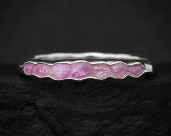 Puede incluir: Un anillo de plata con una hilera de seis pequeñas piedras preciosas rosas en bruto engastadas horizontalmente a lo largo de la banda. La banda de metal tiene un borde ondulado y orgánico que sigue el contorno de cada piedra rosa.