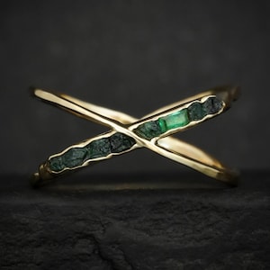 May include: A gold-colored ring with an X-shaped design. The ring is adorned with dark green and emerald-colored gemstones. The gemstones are set in a textured band, creating a unique and elegant look. The ring is displayed against a dark background.