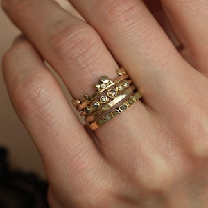 May include: A stack of four gold rings worn on a finger. The top ring features a crown-like design with small gold spheres. The second ring has multiple small, round, dark-colored stones. The third ring is a plain, hammered gold band. The bottom ring is a flat gold band set with a row of small, square-cut, olive-green stones.