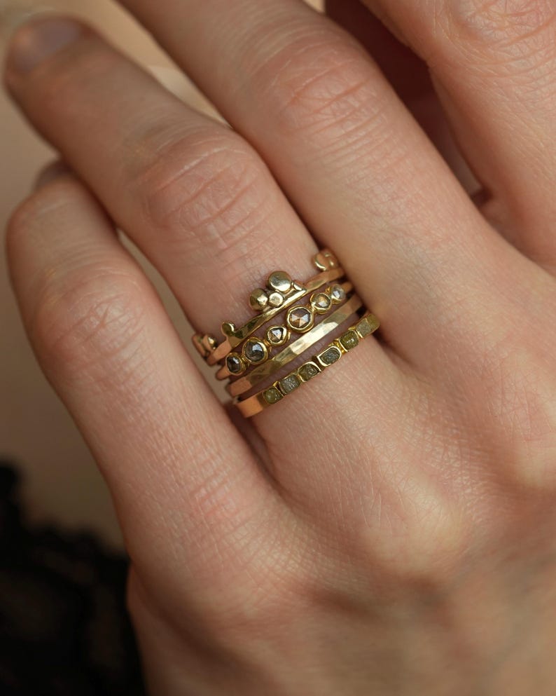 May include: A stack of four gold rings worn on a finger. The top ring features a crown-like design with small gold spheres. The second ring has multiple small, round, dark-colored stones. The third ring is a plain, hammered gold band. The bottom ring is a flat gold band set with a row of small, square-cut, olive-green stones.