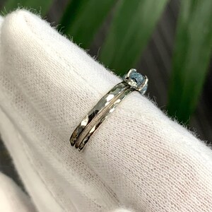 May include: A silver-toned ring featuring a hammered metal band with a textured surface. The ring is set with a single, small, round, light blue stone held in place by a prong setting. The band has a distinct, reflective, and uneven finish.