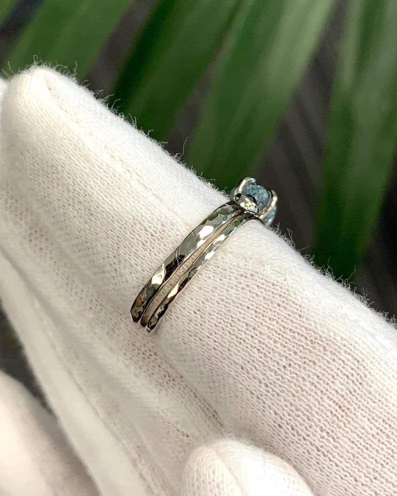 May include: A silver-toned ring featuring a hammered metal band with a textured surface. The ring is set with a single, small, round, light blue stone held in place by a prong setting. The band has a distinct, reflective, and uneven finish.