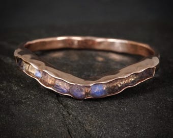 Puede incluir: Un anillo curvo de oro rosa con una textura martillada a lo largo de la banda. El anillo está engastado con una serie de pequeñas piedras de luna azules iridiscentes y redondas que siguen el contorno en forma de V de la banda metálica.