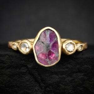 May include: A gold ring with a central, irregular purple gemstone and three smaller, clear gemstones. The ring's design is set against a dark backdrop, accentuating the gold and gemstone colours. The ring is suitable for everyday wear.