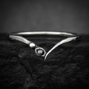 Puede incluir: Un anillo plateado con un diseño curvo único. El anillo presenta una pequeña gema redonda engastada en el centro de la curva. La banda tiene una textura martillada, lo que se suma a su apariencia artesanal. El anillo está sobre un fondo oscuro.