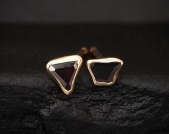 May include: A pair of stud earrings featuring asymmetrical, triangular-shaped dark stones set in polished gold metal frames. The stones have faceted surfaces that catch the light, and the earrings are secured with standard metal posts on the back.