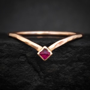 Op de afbeelding: Een goudkleurige ring met een V-vormig ontwerp. De ring heeft een vierkante, robijnrode edelsteen in het midden. De band heeft een gehamerde textuur, wat een unieke touch aan de sieraden geeft.