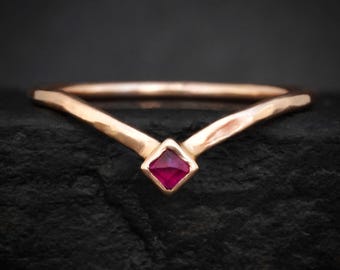 Puede incluir: Un anillo en forma de V fabricado en metal de tono rosado con una textura martillada. El centro de la banda presenta una única piedra preciosa de rubí rojo intenso, de corte cuadrado, engastada en una montura de cuatro garras en la punta.