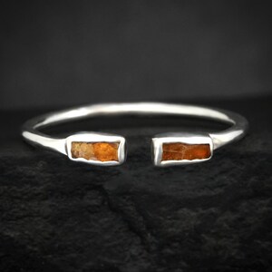 Può includere: Un bracciale aperto color argento con due inserti rettangolari in pietra color ambra. Il bracciale ha un design semplice e moderno, con uno spazio tra le due incastonature di pietre. Lo sfondo è una superficie scura e strutturata.