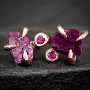 May include: A pair of stud earrings featuring raw, deep magenta-colored crystals set in rose gold metal. Each earring displays a large, irregular, uncut gemstone held by metal prongs, accompanied by a smaller, round, faceted magenta stone.