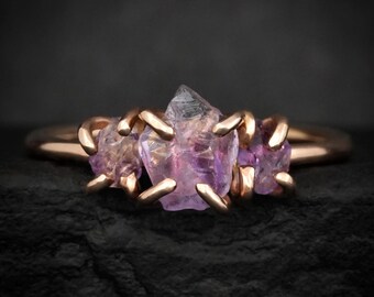 May include: A ring featuring three raw, purple amethyst crystals set in a polished rose gold band. The central amethyst is the largest, flanked by two smaller crystals, all secured by individual rose gold prongs that wrap around the stones.