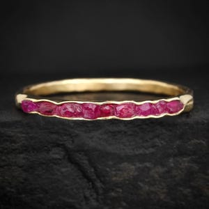 May include: A gold ring with a line of raw ruby gemstones. The ring's band has a textured, hammered look, and the rubies are a deep red colour. The ring is set against a dark background.