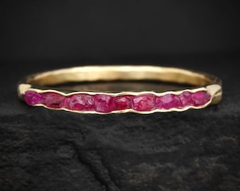 Puede incluir: Un anillo de banda fina de oro con una hilera continua de pequeñas piedras preciosas de rubí en bruto, de color rojo rosado intenso, engastadas horizontalmente en la parte frontal. La banda metálica tiene un acabado liso y pulido y texturas.