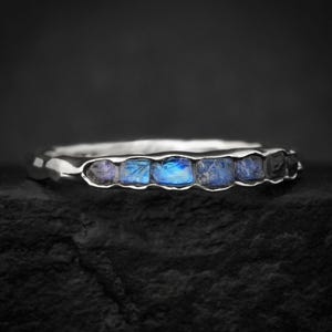 May include: A silver ring featuring a row of six small, rough-cut blue moonstones set into the band. The metal band has a textured, organic appearance with a polished finish, showcasing the iridescent blue flashes of the embedded gemstones.