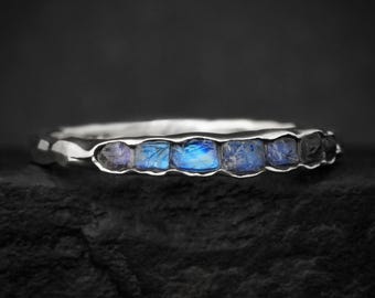 Puede incluir: Un anillo de plata con una hilera de seis pequeñas piedras de luna azules de corte irregular engastadas en la banda. El anillo de metal tiene un aspecto texturizado y orgánico con un acabado pulido, mostrando los destellos azules iridiscentes.