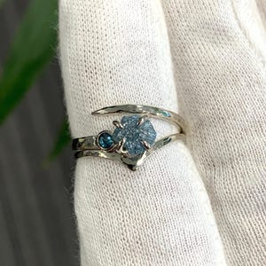 May include: A silver-toned bypass ring featuring a textured, hammered metal band. The ring is set with a central, rough-cut, light blue crystal and a smaller, round, dark blue faceted gemstone positioned next to the larger stone in a prong setting.