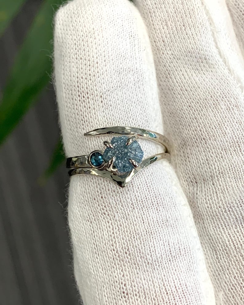 May include: A silver-toned bypass ring featuring a textured, hammered metal band. The ring is set with a central, rough-cut, light blue crystal and a smaller, round, dark blue faceted gemstone positioned next to the larger stone in a prong setting.