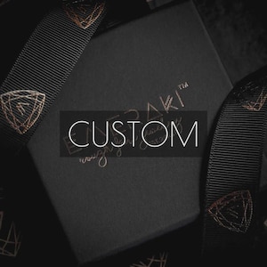 May include: A square black gift box with the word CUSTOM printed in white across the center. The box features a black ribbon with a repeating copper-colored geometric logo pattern. The brand name is embossed in copper on the box lid surface.