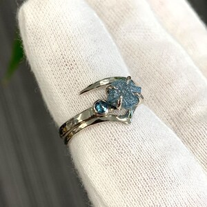 May include: A silver-toned ring featuring a rough, textured blue gemstone set in a prong setting, accompanied by a smaller, round, faceted blue gemstone on a hammered metal band. The ring has a curved, open design that wraps around the finger.