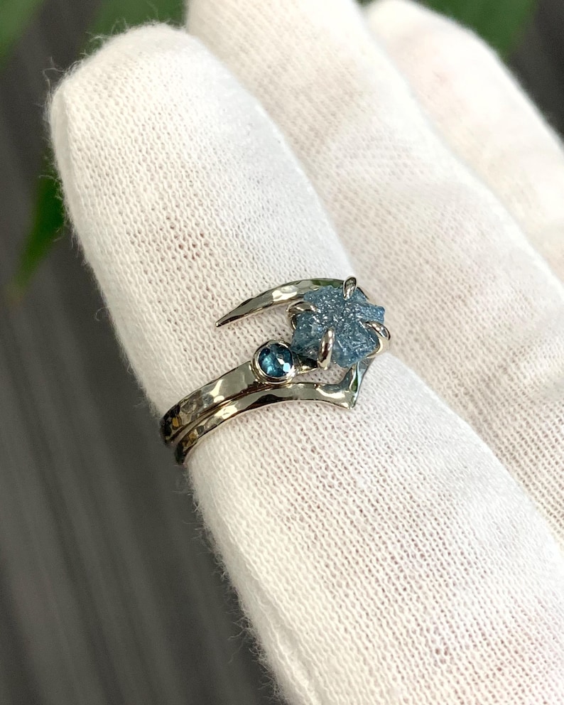 May include: A silver-toned ring featuring a rough, textured blue gemstone set in a prong setting, accompanied by a smaller, round, faceted blue gemstone on a hammered metal band. The ring has a curved, open design that wraps around the finger.