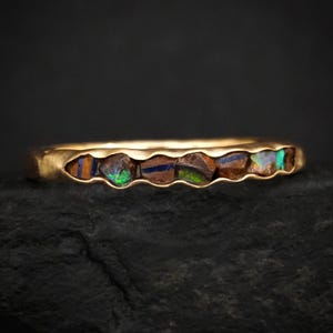 Könnte beinhalten: Ein Goldring mit sechs eingelassenen, rohen Opalsteinen. Die Steine zeigen dunkelbraune, schwarze und tiefblaue Grundfarben mit leuchtenden grünen und blauen Irisierungen. Der Metallring hat eine glatte, polierte Goldoberfläche.