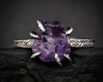 May include: A ring featuring a raw, deep purple amethyst gemstone held by four textured silver prongs. The band is crafted from oxidized silver and displays a repeating, etched herringbone pattern that wraps around the entire circumference.