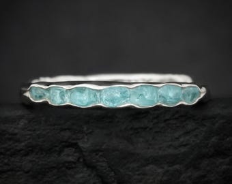 May include: A silver ring featuring a row of seven small, light blue, rough-cut gemstones set horizontally along the band. The metal band has a textured, organic appearance with a polished finish, showcasing the natural shape of the stones.