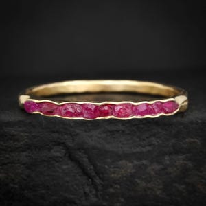 Può includere: Un anello d'oro con una fila di vibranti gemme di rubino grezzo. La fascia ha una texture martellata e i rubini sono di un rosso intenso e ricco. L'anello è ambientato su uno sfondo scuro.