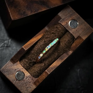 Puede incluir: Un anillo de oro con una gema de ópalo colorido se muestra dentro de una caja de madera con un interior marrón oscuro. El anillo se encuentra sobre un fondo marrón oscuro texturizado. La caja está abierta, revelando los detalles del anillo.