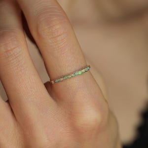 Peut inclure: Une fine bague ornée d'une rangée de petits opales iridescentes serties dans un fin anneau argenté. Les opales présentent des couleurs variées, notamment du vert, du blanc et des touches d'orange. La bague est portée sur un doigt.